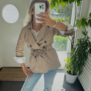 Beige trenchcoat med bälte och knappar - Snygg beige trenchcoat med klassisk krage, dubbelknäppning och knytskärp i midjan. Jackan har långa ärmar och svarta knappar framtill. Perfekt för dig som gillar stilrena och tidlösa plagg med cool siluett. L men passar mig bra som brukar bära s / m