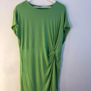 Gina tricot grön klänning  - Grön klänning med en knutdetalj på sidan. Klänningen har en avslappnad passform och korta ärmar. Storlek XL. Fint skick, en liten blekt fläck på ryggen, se sista bild. Inget som syns så mycket