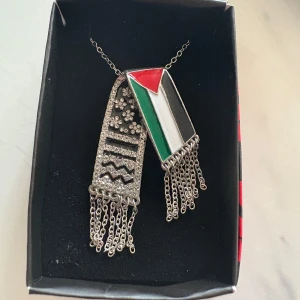 Palestina halsband med kedjor och stenar - Unikt halsband med två hängande berlocker, en med Palestinas flagga i rött, grönt, svart och vitt, och en dekorerad med glittrande stenar och mönster. Båda har kedjefransar längst ner. Finns i både silver- och guldfärgad variant.