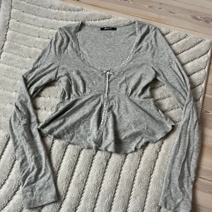 Grå topp med spets - Supersöt grå topp från Gina Tricot med långa ärmar, spetsdetaljer längs kanterna och en slits framtill. Använd 1 gång 
