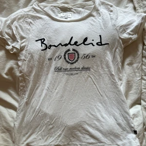 Vit t-shirt från Bondelid med tryck - Vit t-shirt från Bondelid med svart logotyp-tryck och emblem framtill. Klassisk rund halsringning och korta ärmar. Skön och lätt i bomull, perfekt till jeans eller shorts. Snygg och enkel stil med en touch av preppy känsla.❤️🥰