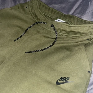 Olivgröna mjukisbyxor från Nike M - Snygga olivgröna mjukisbyxor från Nike med svart logga på benet. Byxorna har dragsko i midjan med svarta och vita snören samt praktiska fickor med svarta dragkedjor. Perfekta för chill eller träning, riktigt sköna och stilrena. pris ej hugget!