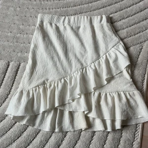 Vit volangkjol med struktur - Supersöt vit kjol med volangdetaljer och strukturerat tyg. Kjolen har en asymmetrisk volang framtill och resår i midjan för skön passform. Det finns en innerkjol som gör att den inte blir genomskinlig