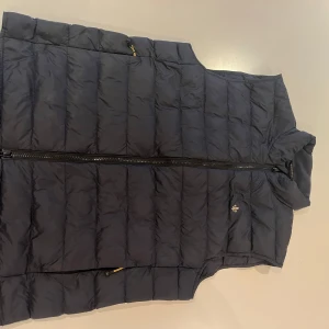 Mörkblå dunväst från Morris - Snygg mörkblå dunväst från Morris med quiltad design och dragkedja framtill. Västen har ståkrage och diskret logga på bröstet. Perfekt för lager-på-lager och håller dig varm under kyliga dagar.nypris 2100, knappt använd, pris kan diskuteras 