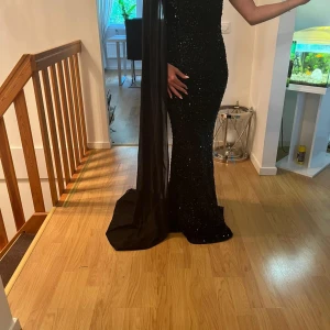 Svart glittrig långklänning oneshoulder - Supersnygg svart långklänning med glittriga paljetter och oneshoulder-design. Klänningen har en lång, elegant släp på ena axeln och sitter tight för en riktigt snygg siluett. Perfekt för dig som vill sticka ut och glänsa på bal eller fest.