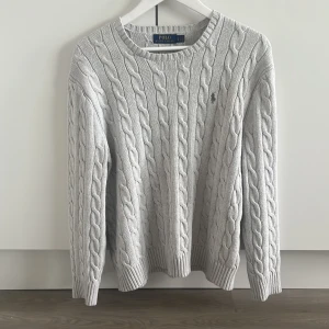 Ljusgrå kabelstickad tröja Polo Ralph Lauren - Kabelstickad tröja från Polo Ralph Lauren i ljusgrått med klassisk rund hals och diskret broderad logga på bröstet. Tröjan är långärmad och har ribbade muddar vid ärmslut och nederkant. Perfekt för lager-på-lager under hösten och vintern. Den har en liten rosa fläck på framsidan men det synns kanppast när man har på sig den. 