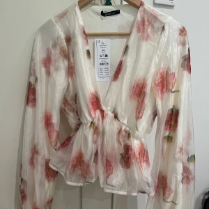 Blommig peplumblus från Gina Tricot - Superfin blommig peplumblus från Gina Tricot i transparent, glansigt material. Blusen är vit med rosa och röda blommor, har v-ringning, långa puffiga ärmar och markerad midja. Perfekt för vår och sommar när du vill känna dig extra söt.