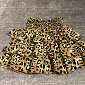 Leopardmönstrad volangkjol S-M - Superfin kort volangkjol med leopardmönster i beige, svart och brunt. Kjolen har bred smock i midjan för skön passform och flera lager volanger som ger en snygg och lekfull look. Perfekt för dig som vill sticka ut lite extra.