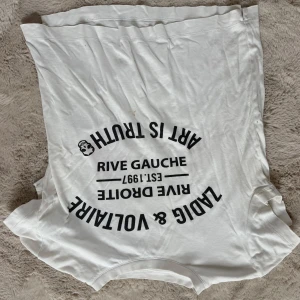 Vit t-shirt med tryck Rive Gauche - Vit t-shirt med svart tryck framtill där det står 'Rive Gauche', 'Art is Truth', 'Zadig & Voltaire' och 'Est. 1997'. Klassisk rund halsringning och korta ärmar. Skön och avslappnad passform, perfekt till jeans eller shorts.