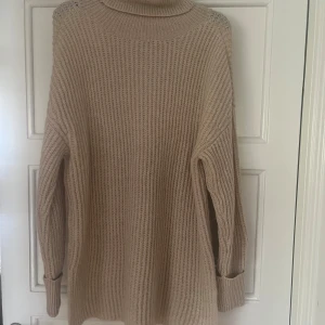 Beige stickad poloklänning i storlek L - Mysig beige stickad poloklänning med ribbstickning och lång ärm. Klänningen har en hög krage och avslappnad passform, perfekt för kyliga dagar. 