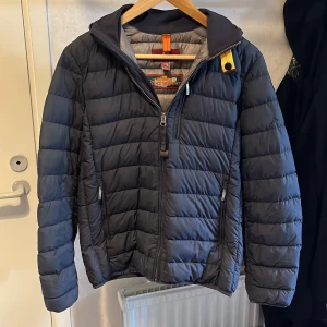 Mörkblå pufferjacka från Parajumpers - Säljer en mörkblå pufferjacka från Parajumpers i storlek Y-M. Jackan är quiltad med horisontella sömmar, har dragkedja framtill och två sidofickor med dragkedja. Detaljer som orange och gul hängare ger en cool touch. Perfekt för kyliga dagar. Den har också ett jätte litet hål som knappt syns