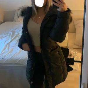 Hollies vinterjacka  - Säljer en svart lång dunjacka från Hollies i modellen Livingo med stor fluffig äkta päls på luvan. Jackan har dragkedja, knappar och flera fickor. Snygg passform och perfekt för kalla dagar. Pälsen är naturfärgad med svarta inslag och jackan har justerbar midja. Använd 2 vintrar, det finns inga defekter på jackan. Storlek 32, passar på S och M Nypris: 5000kr 