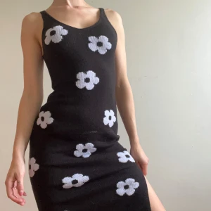 Svart blommig stickad långklänning - Svart stickad långklänning med vita blommor över hela klänningen. Ärmlös modell med v-ringning och slits på sidan. Sitter figurnära och har en mjuk, stretchig känsla. Perfekt för dig som gillar retrovibbar och vill sticka ut.