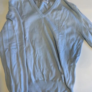 Ljusblå stickad tröja från Wolsey - Ljusblå stickad tröja från Wolsey med v-ringning och ribbade muddar. Klassisk och enkel design, perfekt för lager-på-lager. Tillverkad i Skottland och har en diskret broderad logga nertill. Skön och mjuk känsla.