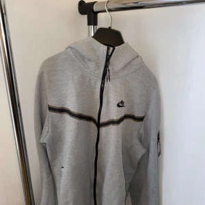 Grå Nike hoodie med dragkedja - Säljer en grå hoodie från Nike med hel dragkedja och svart logga på bröstet. Tröjan har huva, svarta detaljer och en praktisk ficka med dragkedja på ärmen. Perfekt för chill dagar eller när du vill ha en sportig look.