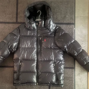 Svart glansig vinter jacka Ralph Lauren L - Säljer en svart glansig dunjacka från Ralph Lauren med huva och dragkedjor. Jackan har en klassisk pufferstil, tre fickor med dragkedja och ikonisk röd logga på bröstet. Perfekt för kalla dagar och riktigt snygg streetstil, säljer den pågrund av att den är för stor för mig. Om du har några frågor skriv gärna😊