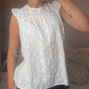 Vit blommig blus med volangärm från pigalle - Säljer en söt vit blus med diskret blommigt mönster och volangdetaljer vid ärmslut. Blusen är ärmlös och har knytning i nacken som ger en fin öppen rygg. Perfekt för varma dagar och enkel att styla till jeans eller kjol.