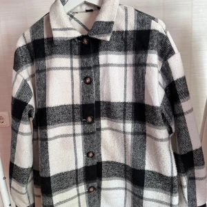 Rutig Skjortajacka - Snygg overshirt i svartvitt rutigt mönster med klassisk krage och knäppning framtill. Jackan är gjord i en mjuk ullmix och har långa ärmar. Perfekt för lager-på-lager och ger en cool vibe till din outfit.