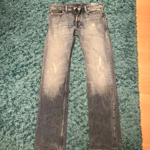 Diesel jeans - Snygga blå jeans från Diesel med slitna detaljer och klassisk femficksdesign. Jeansen har raka ben och en normal passform. Tydliga slitningar framtill och på bakfickorna ger en cool, använd look. Perfekta till sneakers och hoodie.