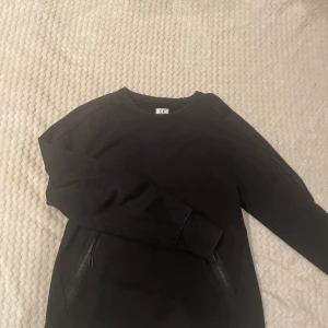Svart sweatshirt från C.P. Company - Svart sweatshirt från C.P. Company med klassisk rund hals, långa ärmar och två dragkedjefickor framtill. Ärmen har den ikoniska C.P. Company-linsen och en extra ficka med dragkedja. Perfekt för en stilren och sportig look. Ny pris  200kr