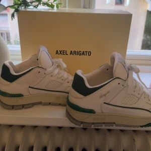 AXEL ARIGATO AREA LO SNEAKERS vita med gröna detaljer - Snygga vita sneakers från Axel Arigato i storlek 39. Original pris 3450kr. Skorna har gröna detaljer och en chunky beige sula. Klassisk modell med snörning och stilren design. Tillverkade i skinn och återvunnen polyester. Handgjorda i Portugal. Perfekta för dig som gillar trendiga och bekväma sneakers.