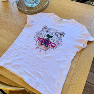 Vit Kenzo t-shirt med tigertryck - Vit t-shirt från Kenzo med ikoniskt tigertryck i rosa, svart och beige på bröstet samt texten 'Kenzo Paris' i rosa. Klassisk rund hals och korta ärmar. Tillverkad i mjuk bomull för en skön känsla. Perfekt statement-plagg för dig som gillar streetstyle.