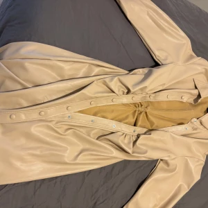 Beige lång skinnjacka med knappar - Säljer en beige lång skinnjacka med snygga tryckknappar längs hela framsidan. Jackan har lång ärm och ett stilrent, minimalistiskt utseende. Materialet är mjukt och har en lätt glansig finish. Perfekt för dig som gillar clean och modern stil.