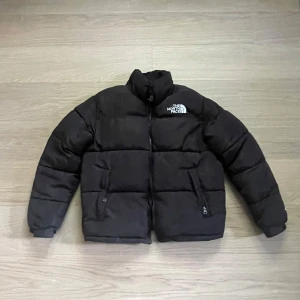 Svart pufferjacka från The North Face - Säljer en svart pufferjacka från The North Face med hög krage och dragkedja framtill. Jackan har två fickor med dragkedja och logga på både bröstet och ryggen. Perfekt för kalla dagar och riktigt snygg till streetwear-stilen.