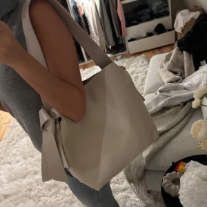 Beige axelväska med rosett i skinn - Stilren beige axelväska i slätt skinn med bred axelrem och snygg rosettdetalj på sidan. Väskan har en minimalistisk design och rymmer allt du behöver till vardagen. I nyskick!
