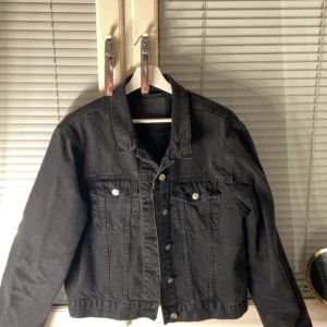 Svart jeansjacka med silvriga knappar - Säljer en svart jeansjacka med klassisk passform och silvriga knappar framtill. Jackan har två bröstfickor med lock och knapp, samt snygga sömdetaljer. Perfekt att slänga över en hoodie eller t-shirt för en chill look.