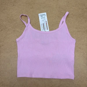 Rosa ribbad linnetopp från B Sister - Säljer en ljusrosa ribbad linnetopp från B Sister. Toppen är croppad och har smala axelband, perfekt för varma dagar. Tillverkad i mjuk bomull med stretch för en bekväm passform. Enkel och stilren design som är lätt att matcha med jeans eller kjol.