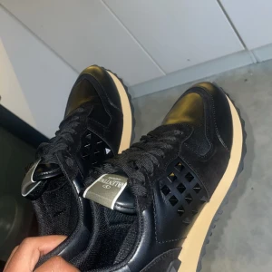 Valentino Garavani svarta sneakers - Snygga svarta sneakers från Valentino Garavani med coola nitar på sidorna och beige sula. Skorna har snörning och är tillverkade i Italiea och mocka för en lyxig känsla. Perfekta för dig som vill sticka ut med stilrena detaljer. Priset kan diskuteras 
