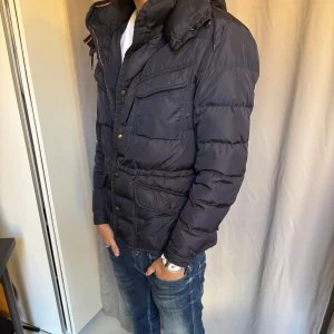 Mörkblå Moncler jacka - Snygg mörkblå jacka från Moncler. Snygg luva med dragsko och detaljer i guld på knapparna. Jackan har lång ärm och en klassisk quiltad look som ger extra värme. Jackan har en nål på dragkedjan men är perfekt för kalla dagar och stilren till jeans. Hör gärna av er vid frågor! .🙌🙌