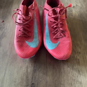 Nike Mercurial rosa fotbollsskor - Säljer ett par Nike Mercurial fotbollsskor i en snygg rosa färg med ljusblå detaljer och stora Nike-loggor. Skorna har snörning och är tillverkade i syntetmaterial för lätt känsla och bra grepp på planen. Perfekta för dig som vill sticka ut på fotbollsplanen.