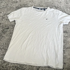 Vit t-shirt från GANT i bomull - Vit klassisk t-shirt från GANT med rund halsringning och diskret logga på bröstet. Tillverkad i mjuk bomull och har en enkel, stilren design med korta ärmar. Perfekt för en clean och avslappnad look.