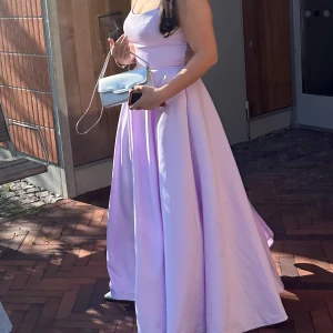 Ljuslila lång/balklänning med smala axelband - Superfin långklänning i ljuslila med smala axelband och korsad rygg. Den är i storlek S men går att justera, jag brukar ha L och den kan passa alla. Jag är 175cm lång och den gick precis ner i marken. Klänningen har en figurnära överdel och ett utsvängt, flowy kjolparti. Materialet är glansigt och känns lyxigt, perfekt för bal eller student. Stängs med dragkedja i ryggen.