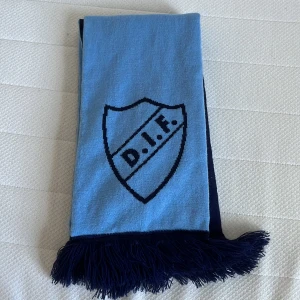 Djurgården halsduk blå och marinblå, aldrig använd  - Säljer en Djurgården halsduk i blå och marinblå färg med DIF-logga och texten 'Stockholmslaget' samt 'Djurgården' på sidorna. Halsduken har fransar i ändarna och är perfekt för att visa ditt stöd på matchen. Materialet är stickat och mjukt.