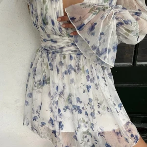 Blommig vit och blå kortklänning - Superfin kortklänning i vitt med blått blommönster och gröna detaljer. Klänningen har v-ringning, markerad midja och vida, långa ärmar med volang vid ärmslut. Tunt, luftigt material som ger en romantisk vibe och flowig siluett. Storlek 44 men passar L🤍