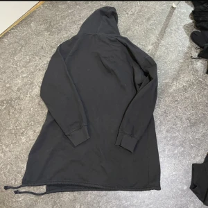 Svart lång hoodie med snörning - Svart hoodie med lång passform och huva. Tröjan har långa ärmar och snörning längst ner i kanten för att justera stilen. Tillverkad i mjukt bomullsmaterial, perfekt för chill dagar. Enkel och clean design utan tryck eller mönster.