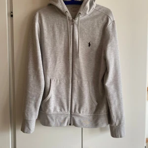 Grå hoodie från Polo Ralph Lauren - Grå hoodie från Polo Ralph Lauren med dragkedja, huva och snörning. Klassisk logga broderad på bröstet och två fickor framtill. Tillverkad i mjuk bomullsmix, perfekt för chill dagar. Snygg och enkel design som funkar till allt. Priset är inte fast👍