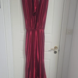 Vinröd långklänning i satin - Snygg vinröd långklänning i glansigt satinmaterial med djup v-ringning och ärmlös design. Perfekt för dig som vill sticka ut med en elegant och stilren look. Klänningen har markerad midja och ett vackert fall.