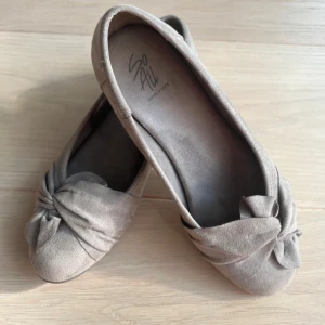 Beige/gråa ballerinaskor - Snygga beige ballerinaskor från So All i mjuk mocka-look. Skorna har en rund tå och en dekorativ rosett framtill som ger en söt touch. Platt sula gör dem bekväma för hela dagen. Perfekta att matcha med både jeans och klänning. Bara att dm:a vid frågor eller funderingar!💕Pris kan diskuteras💕