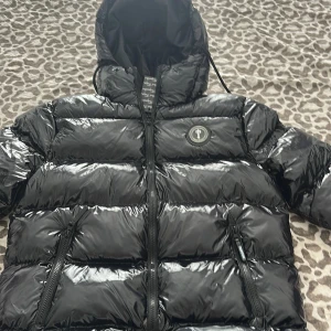 Svart pufferjacka från Trapstar - Säljer en svart, glansig pufferjacka från Trapstar med huva och tydlig logga på bröstet. Jackan har dragkedja framtill och två snedställda fickor med dragkedja. Perfekt för kalla dagar och ger en riktigt streetig vibe.