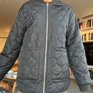 Svart quiltad bomberjacka - Quiltad svart bomberjacka med rundad hals, två stora fickor framtill och dragkedja hela vägen. Jackan har ett snyggt mönster i tyget och ribbade muddar vid ärmslut och hals. Perfekt för dig som gillar stilren och enkel design.