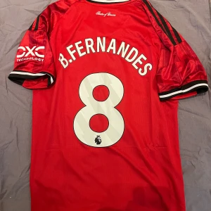 Manchester United BrunoFernandes tröja - Röd Manchester United matchtröja från Adidas med B. Fernandes och nummer 8 på ryggen. Tröjan har svarta och vita detaljer, klubbmärke på bröstet, sponsorloggor och klassiska Adidas-ränder på axlarna. Tillverkad i lätt och ventilerande material.