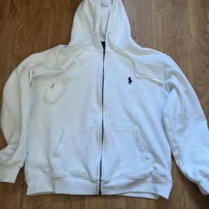Vit hoodie från Polo Ralph Lauren - Vit hoodie från Polo Ralph Lauren med dragkedja, huva med snörning och klassisk broderad logga på bröstet. Mjuk bomullskvalitet och känguruficka framtill. Perfekt för en clean och stilren look.