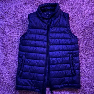 Svart dunväst från Everest 146/152 - Svart dunväst från Everest med horisontella quiltade sömmar och hög krage. Västen har dragkedja framtill och två sidofickor. Materialet är syntet och dun, perfekt för kyliga dagar. Snygg och enkel design med diskret Everest-logga på bröstet. Perfekt till när hösten närmar sig! 🍂 
