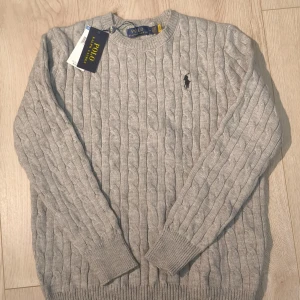 Grå Knit Polo Ralph Lauren M - Kabeltickad Ralph Lauren tröja i grå med den klassiska Polo-loggan på bröstet. Tillverkad i 100% mjuk bomull som sitter bekvämt och håller formen fint. 📏Storlek M, längd 65 cm och bredd 48,5 cm. Oanvänd i fint skick. Meddela för frågor!