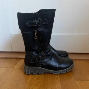Svarta boots med spännen och dragkedja 35 - Tuffa svarta boots i skinnimitation med grov sula, dragkedja på sidan och snygga spänndetaljer. Ovankant i mjukt teddyfoder för extra värme i storlek 35