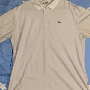 Vit pikétröja från Lacoste - Vit pikétröja från Lacoste med klassisk krage och korta ärmar. Tröjan har den ikoniska broderade krokodilloggan på bröstet och knäppning med två knappar. Materialet är mjuk bomull som andas och passar perfekt till en chill stil.